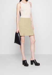 Top sans manches rose clair, jupe jaune avec fentes latérales, mules noires à talons hauts et un sac à main noir. Textures lisses, design moderne.