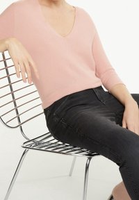 Różowa dziana bluzka z dekoltem w serek, zestawiona z ciemnymi dżinsami z bocznymi szwami, noszona przez osobę siedzącą na chromowanym krzesle z drutów.