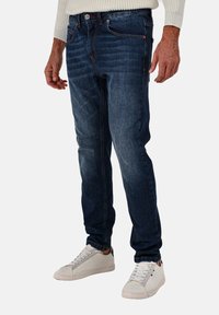 Jeans in denim blu scuro con vestibilità dritta, caratterizzati da un leggero sbiadimento e cuciture arancioni, indossati con sneakers bianche.