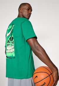 Green cotton T-shirt with a graphic print on the back, featuring a black and green design. Model holds an orange basketball.