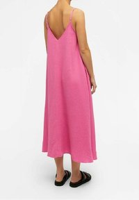 Robe longue rose sans manches avec un dos en V, présentant un tissu texturé et une silhouette fluide, associée à des sandales noires.