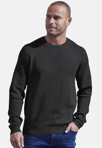 Schwarzer strukturierter Pullover mit geripptem Rundhalsausschnitt und diagonaler Streifenmuster, kombiniert mit blauen Jeans. Das Model steht lässig.