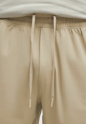 Pantaloni sportivi beige con vita elastica, dotati di lacci e tasche laterali. Texture liscia con dettagli di cucitura minimi.