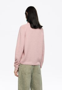 Maglione rosa chiaro lavorato a maglia con scollatura tonda e maniche lunghe, abbinato a pantaloni a gamba larga verde oliva. Texture morbida, vestibilità ampia.