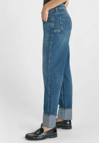 Un jean en denim de couleur bleu moyen, avec une coupe décontractée, des jambes droites, des ourlets retroussés de couleur bleu clair contrastante et des poches latérales. Des mocassins noirs sont portés.