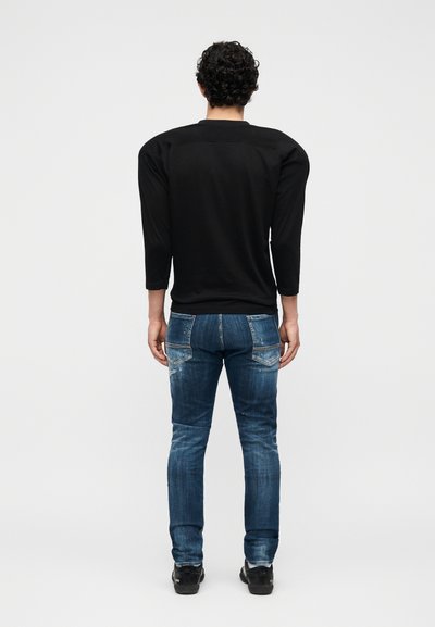 Άνδρας με σγουρά μαλλιά στέκεται με την πλάτη προς το μέρος μας, φοράει μαύρο μακρυμάνικο πουκάμισο, μπλε τζιν και μαύρα παπούτσια σε απλό λευκό φόντο.