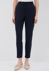 Pantaloni tailored blu navy con vestibilità slim, caratterizzati da una parte anteriore piatta, passanti per la cintura e spacchi alla caviglia. Indossati con scarpe con tacco beige.