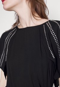 Black top featuring a loose fit with short sleeves. Decorative silver studs along the shoulders. Soft texture and flowing design.