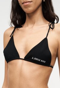 Κοντινό πλάνο γυναίκας που φοράει μαύρο τρίγωνο μπικίνι με λεπτές τιράντες και το κείμενο «A. ROEGE HOVE» στο αριστερό κύπελλο.