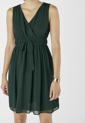 Bez rękawów, ciemnozielona sukienka do kolan z dekoltem w serek, marszczoną talią i paskiem z tkaniny, noszona z złotą bransoletką na prawej ręce.