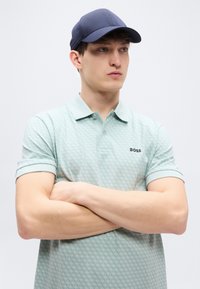 Πόλο μπλούζα σε μέντα πράσινο με γεωμετρικό μοτίβο, με γιακά και κοντά μανίκια που είναι γυρισμένα. Συνδυασμένη με καπέλο σε ναυτικό μπλε χρώμα.