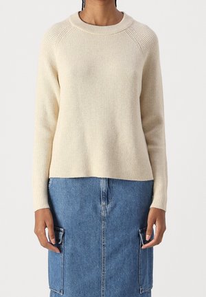 Personne portant un pull crème à manches longues côtelé associé à une jupe en jean bleue avec deux poches à rabat à l'avant.