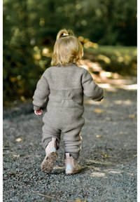 Šedý overal z fleecu s žebrovanými manžetami, doplněný světle růžovými kotníkovými botami. Dítě kráčí po štěrkové cestě v přírodním prostředí.