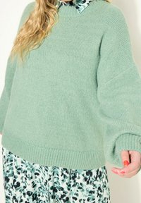 Donna che indossa un maglione lavorato a maglia verde chiaro sopra un vestito collared verde e nero a fantasia, con lunghi capelli biondi mossi e smalto rosso sulle unghie.