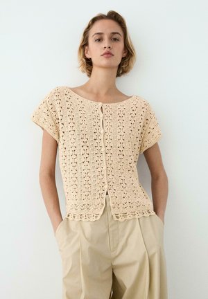 Kobieta ubrana w beżowy, krótkim rękawem, szydełkowany top na guziki, połączony z beżowymi plisowanymi spodniami z wysokim stanem, stojąca na tle jednolitej jasnej ściany.
