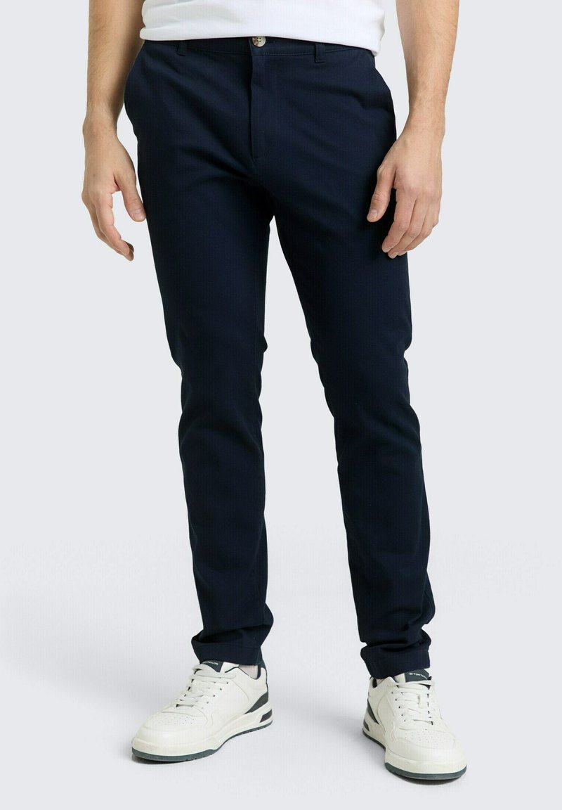 Στενές παντελόνες σε ναυτικό μπλε χρώμα, κατασκευασμένες από απαλό ύφασμα, με στενή εφαρμογή, μπροστινές τσέπες και κούμπωμα με κουμπί, συνδυασμένες με λευκά αθλητικά παπούτσια.