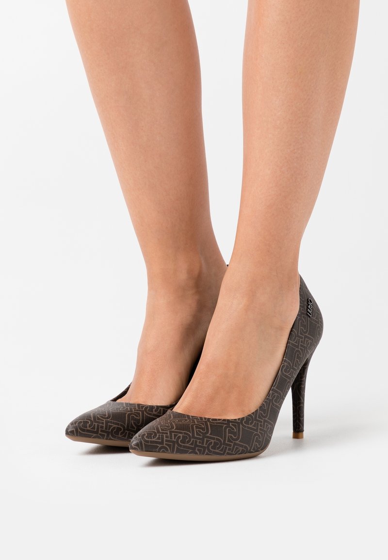 Jambes portant des chaussures à talons hauts marron foncé avec un motif géométrique et un bout pointu, sur fond blanc.