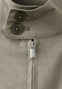 Približna slika ovratnika bež oblačila z dvema temnima gumboma in srebrnim zadrgovanjem, na katerem je vgravirano »NIN9«.