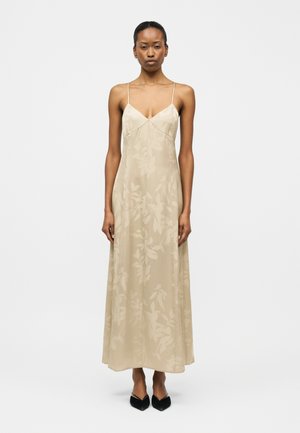 Moteris dėvi smėlio spalvos, be rankovių, iki kulkšnių ilgio suknelę su subtiliu lapų raštu ir plonomis petnešėlėmis, derintą su juodais smailiais bateliais.