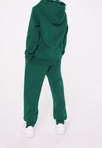 Grön huvtröja och matchande mjukisbyxor, tillverkade av mjukt tyg med en avslappnad passform. Har en huva och elastiska ärmslut, i kombination med vita sneakers.