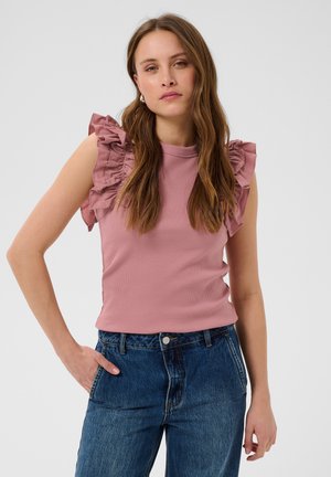 Femme aux longs cheveux bruns porte un haut sans manches rose poudré avec des volants sur les épaules et un jean bleu taille haute, debout devant un fond uni.