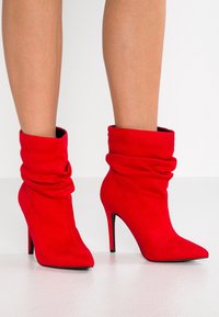 Botas de tobillo de ante rojo con un diseño suelto, punta afilada y alto tacón de aguja, con una textura suave y acentos de suela negra.