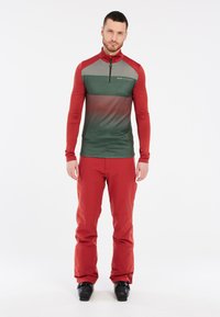 Rood en groen langlopend shirt met geometrische patronen en een halflange ritskraag, gecombineerd met effen rode broek en zwarte skibotten.