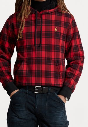 Osoba nosząca czerwoną i czarną bluzę w kratę z czarnymi sznurkami i małym żółtym logo, w połączeniu z ciemnymi dżinsami i czarnym paskiem.