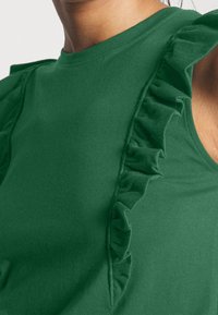 Gros plan d'un haut vert sans manches présentant un détail en tissu froncé vertical le long de l'épaule et de la poitrine.