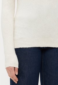 Detailní záběr osoby v krémovém pleteném svetru a tmavě modrých džínách, kde je levá ruka uvolněně položena na boku.