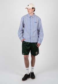 Joven con gorra beige, camisa abotonada con rayas verticales azules y rosas, pantalones cortos verde oscuro, calcetines blancos y zapatos negros, de pie con las manos en los bolsillos.