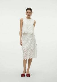 Ensemble en dentelle blanche composé d'un haut à ourlet festonné et d'une jupe midi à motifs ondulés; associé à des sandales à glissière florales rouges.