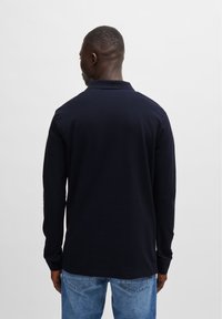 Marineblauwe polo met lange mouwen en een kraag, met een gladde textuur en zijsplitten aan de zoom. G dragen met lichtblauwe jeans.