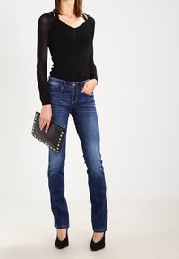 Zwart langemouwen top met een gestructureerd patroon, gecombineerd met blauwe denim jeans. Accessoires zijn een zwarte studs clutch en zwarte hakken.
