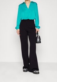 Teal blouse with a V-neckline and gathered sleeves paired with high-waisted black flared pants; model holding a small black handbag.