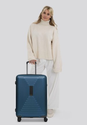 Donna bionda in maglione beige a collo alto e pantaloni bianchi in piedi, mentre tiene il manico di una grande valigia rigida blu con ruote.