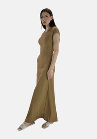 Robe longue et moulante en tissu brun texturé, avec des manches courtes et une fente sur le côté. Assortie à des sandales plates de couleur claire.