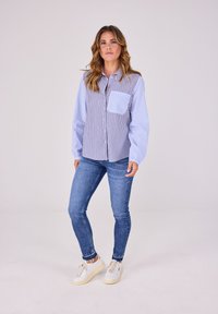 Gestreiftes Button-up-Hemd in Blau und Weiß, mit einer Brusttasche, kombiniert mit eng anliegenden blauen Jeans und weißen Sneakers.