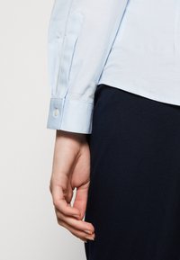 Gros plan sur le bras gauche d’une personne portant une chemise à manches longues bleu clair avec poignets boutonnés et un pantalon sombre sur un fond uni.