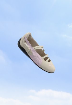 Chaussure beige clair et rose à enfiler avec des bandes élastiques et une semelle noire flottant sur un fond de ciel bleu.