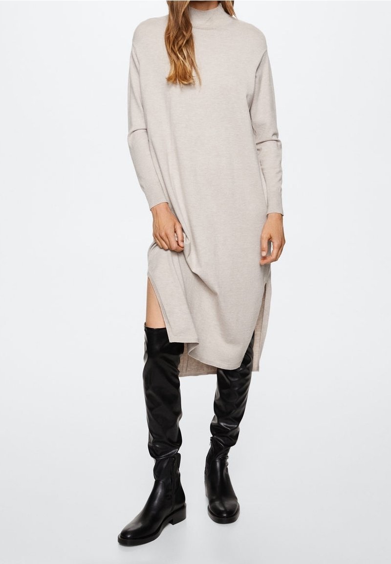 Femme portant une robe midi beige ample à manches longues avec des fentes latérales et des bottes en cuir noires montant jusqu'aux genoux, debout sur fond blanc.