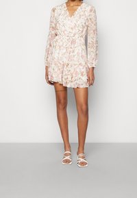 Vestido cruzado floral en rosa claro y crema con mangas largas, dobladillo con volantes y cintura con cordón ajustable. Combinado con sandalias blancas de tiras.