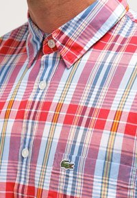 Koszula zapinana na guziki z bawełny w wielokolorową kratkę z czerwonymi, niebieskimi i żółtymi elementami. Zawiera logo Lacoste na piersi.