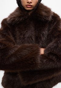 Casaco de pêlo sintético castanho com textura longa e suave, corte oversized e mangas largas. Apresenta um colarinho alto e detalhes mínimos.