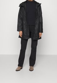 Schwarze wasserdichte Parka-Jacke mit Reißverschluss und Druckknöpfen, ausgestattet mit einer abnehmbaren Kapuze, getragen über einem schwarzen Oberteil und einer schwarzen Hose.