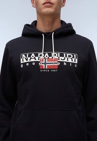 Black hooded sweatshirt with a front pocket, featuring a white logo and a red and blue flag design. Cotton blend fabric, casual fit.