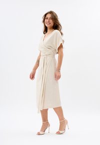 Beige Wickelkleid mit kurzen Ärmeln, Bindegürtel in der Taille und dezentem Blattmuster. Kombiniert mit sandalen mit hohen Absätzen und Riemchen. Glatte Textur.