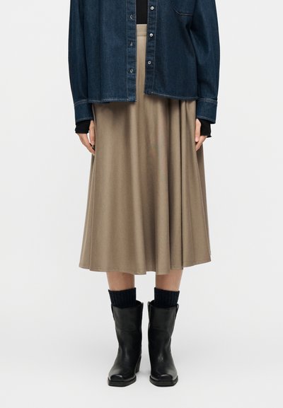 Frau trägt einen beigen Midirock, schwarze knielange Stiefel mit dunklen Socken und eine dunkle Jeansjacke über einem schwarzen Oberteil, steht vor einem weißen Hintergrund.