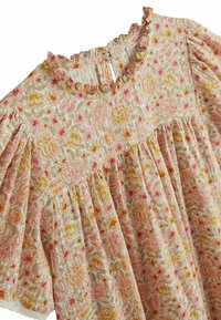 Blouse à motif floral avec un col froncé et des manches courtes bouffantes. Présente un fond beige avec des motifs floraux roses, jaunes et verts.