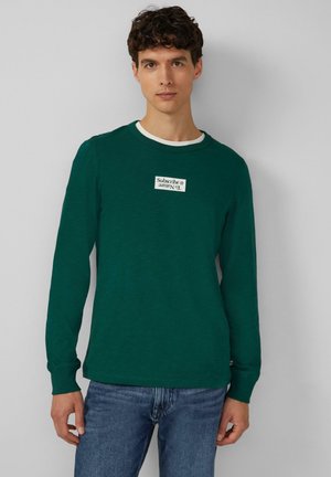 Grünes langärmliges T-Shirt aus weichem Material, mit einem weißen Ausschnitt und einem Aufnäher mit dem Text "Abonnieren" auf der Brust.